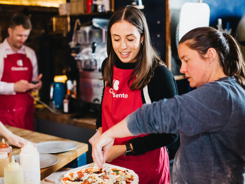 A woman in a blue jumper is showing a woman in a red apron how to make a handmade pizza